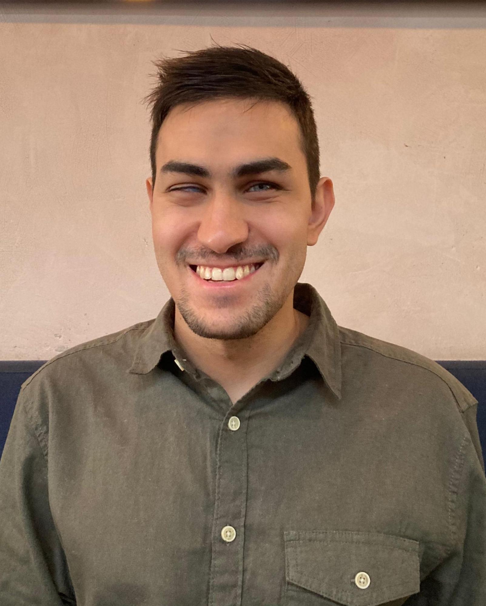 Foto de Gustavo Torniero do peito para cima, sorridente. Ele tem pele clara, cabelos castanho-escuros, olhos azuis e barba curta. Usa uma camisa de botão verde-escura, com bolso no lado esquerdo do peito. Ao fundo, uma parede bege-clara.